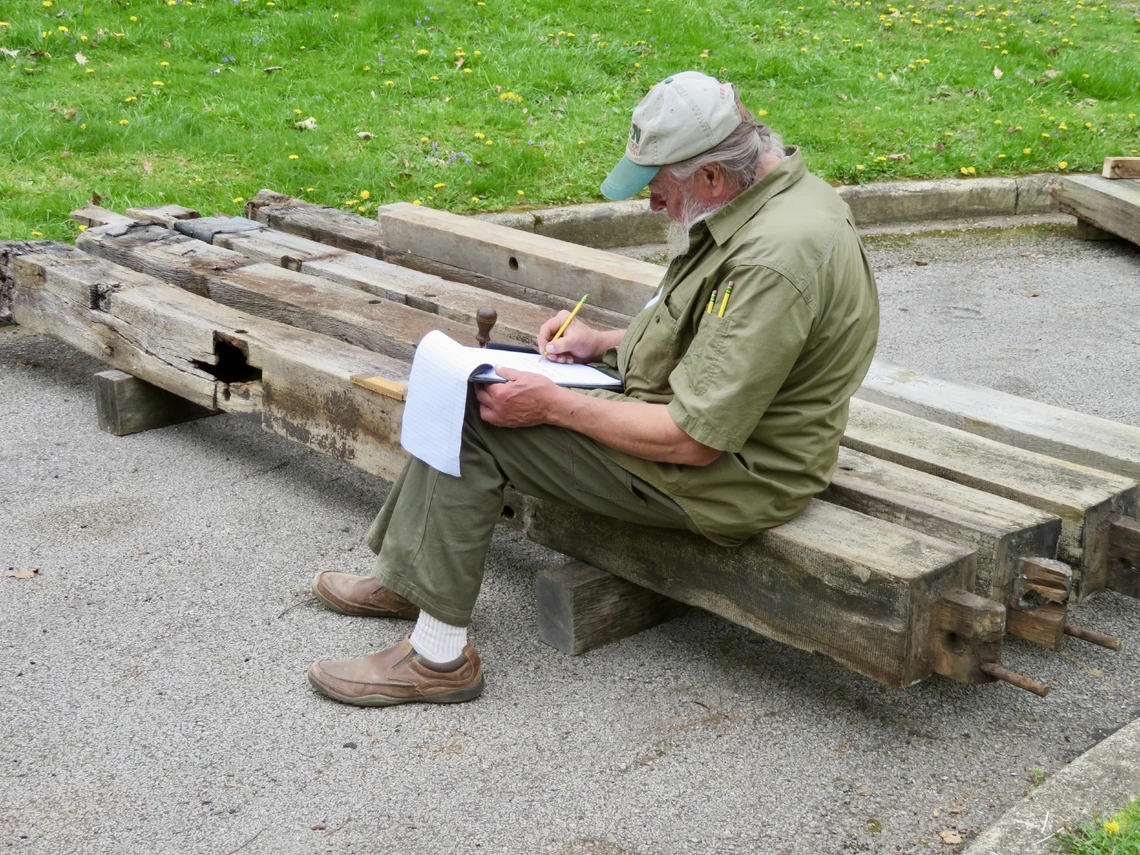 A man with a white beard sits outside on a wooden beam on the ground to write notes on a pad of paper.