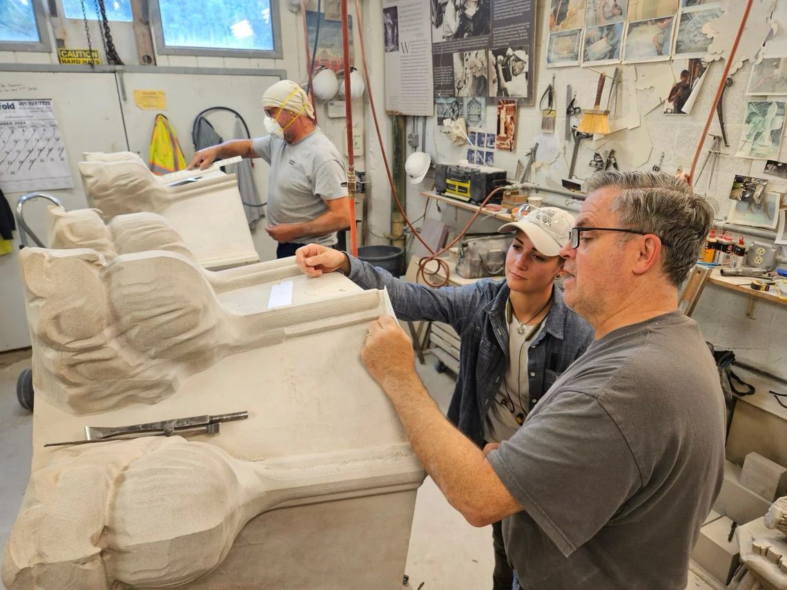 Inside a stone masons’ workshop, a man shows a young woman how to trace the outlines of a cardboard template onto a Gothic-style pinnacle stone in preparation for carving; another man is in the background carving a piece of stone on a workbench.