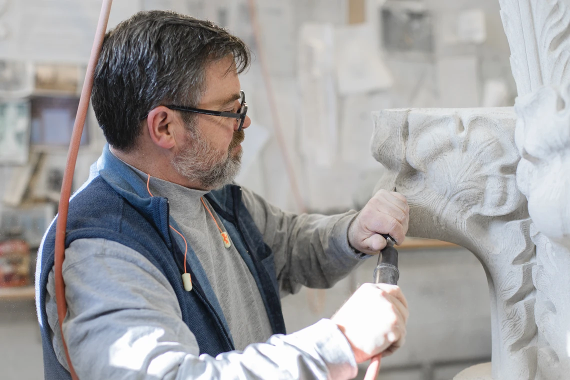 A man in a stone carving workshop uses a metal air hammer and chisel to carve decorative foliage on a Gothic-style limestone final.