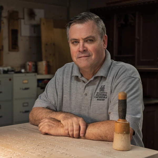 A stone carver sits in his workshop next to a limestone tablet with hand-carved inscriptions on a worktable; his mallet sits on the stone tablet in front of him.