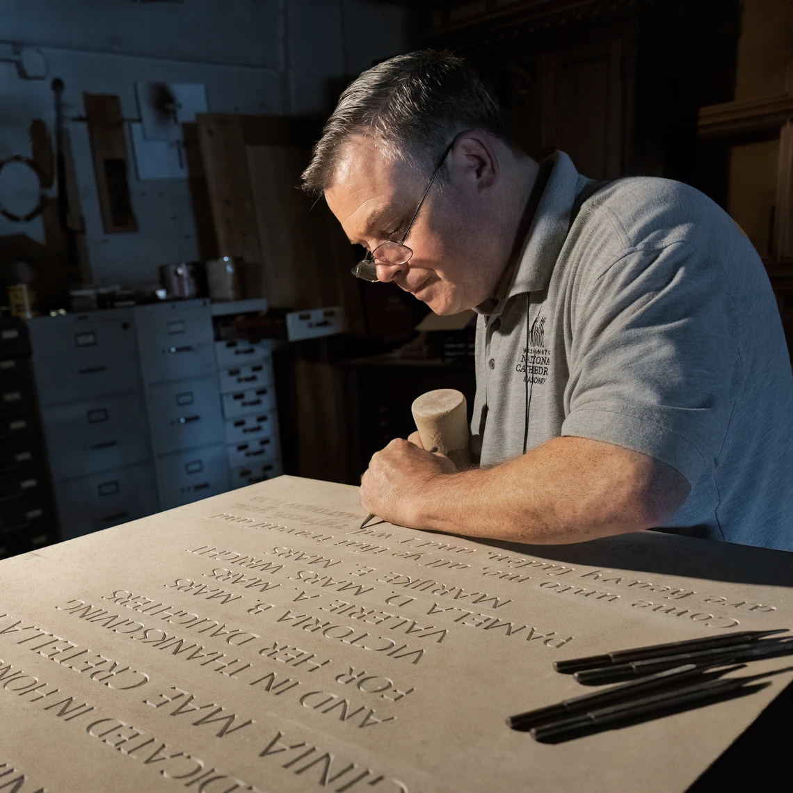 A man in a stone carving workshop uses a mallet and chisel to hand-carve inscriptions into a limestone tablet.