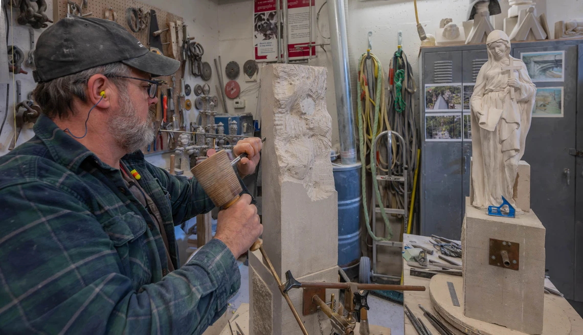 Inside a stone masons’ workshop, a man uses a wooden mallet and chisel to rough out a small statue of a female saint in stone; the plaster model of the saint is on a worktable next to him to serve as a point of reference.