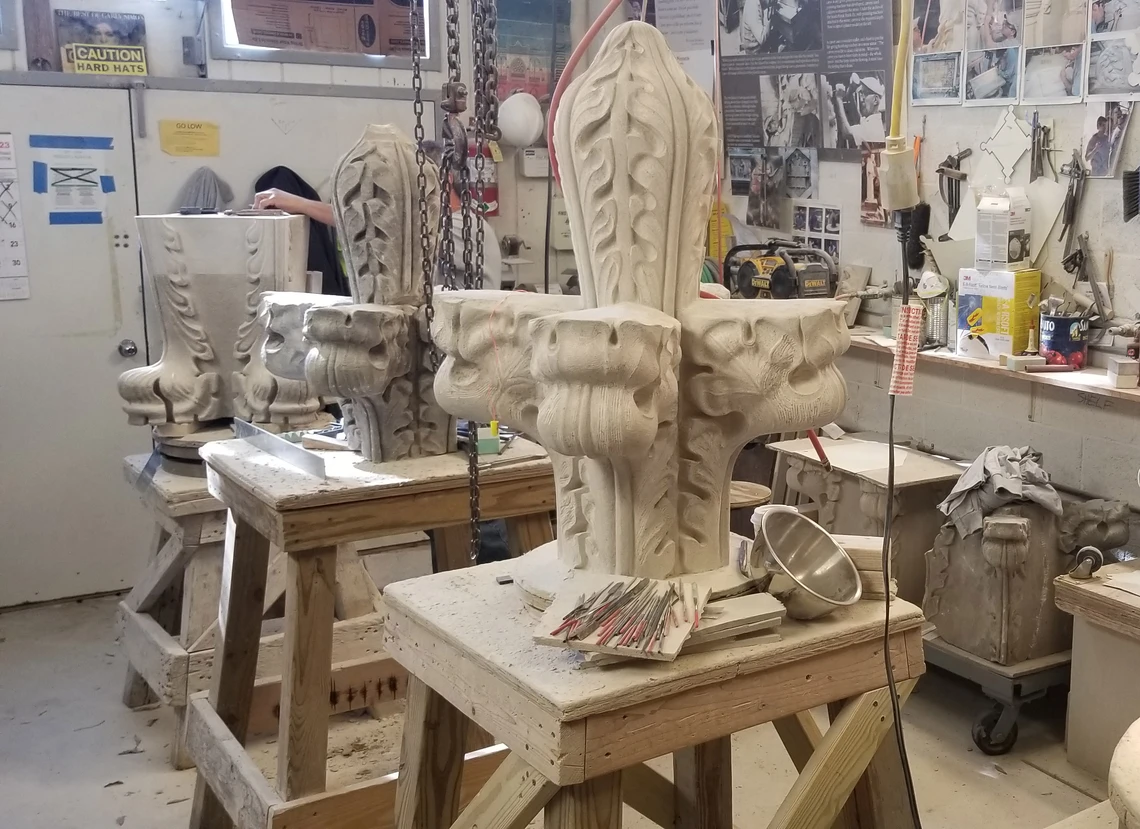 Inside a stone masons’ workshop, two Gothic-style finial stones and a pinnacle stone sit atop sturdy wooden workbenches; various carving tools, architectural drawings, and photographs hang on the wall in the background.