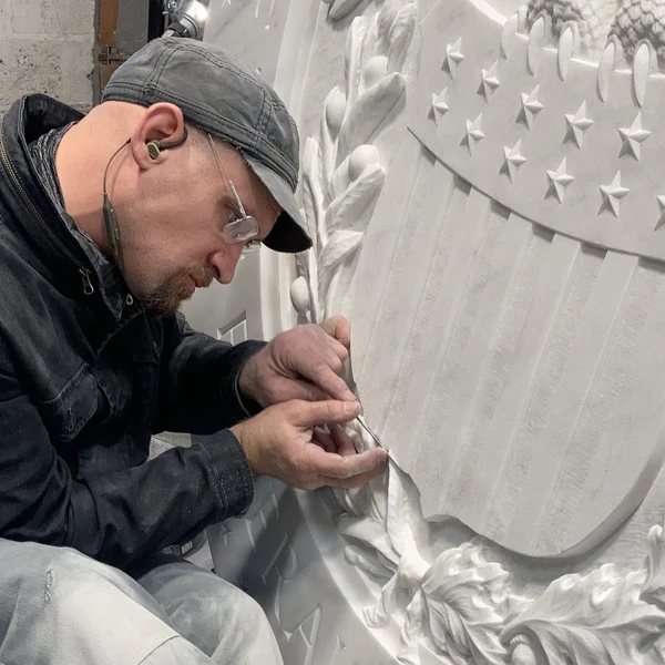 A man in a black cap uses a small metal file to put the finishing touches on a stone sculpture featuring a cluster of oak leaves and a plaque with stars and stripes.