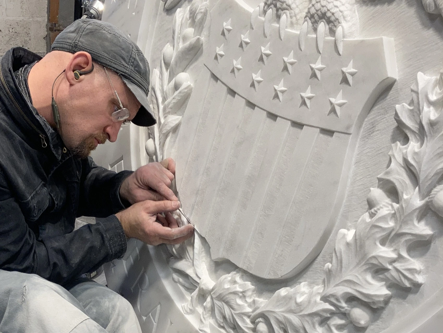 A man in a black cap uses a small metal file to put the finishing touches on a stone sculpture featuring a cluster of oak leaves and a plaque with stars and stripes.