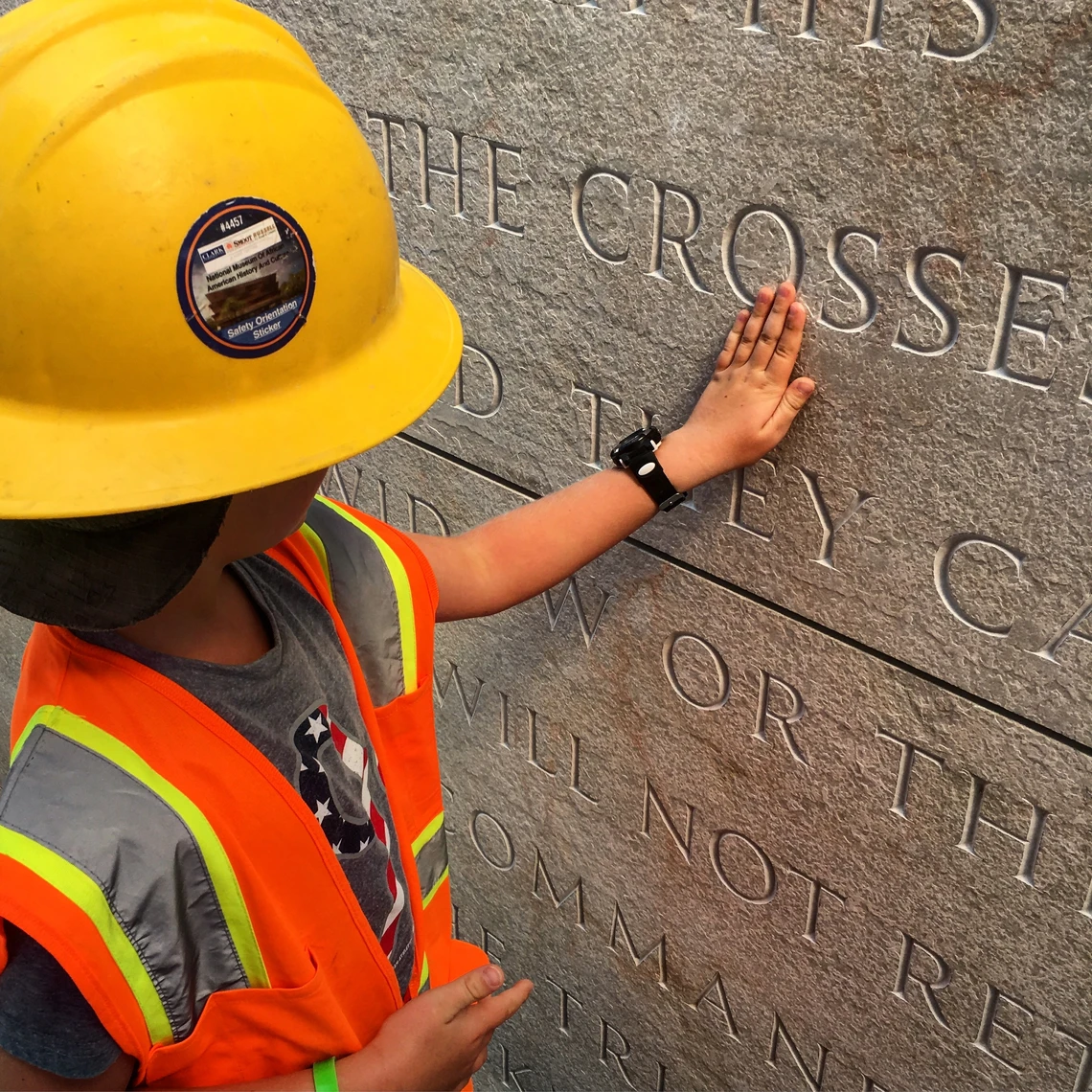 A young boy in a yellow hard hat and orange safety vest touches letters carved in an exterior stone wall.