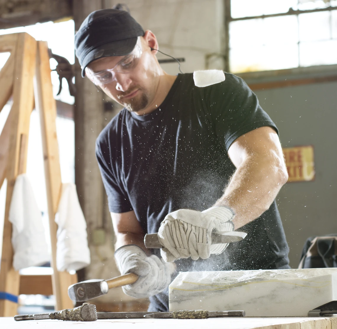 A man in a stone carving studio strikes a piece of marble on a workbench with a steel hammer and thick chisel.