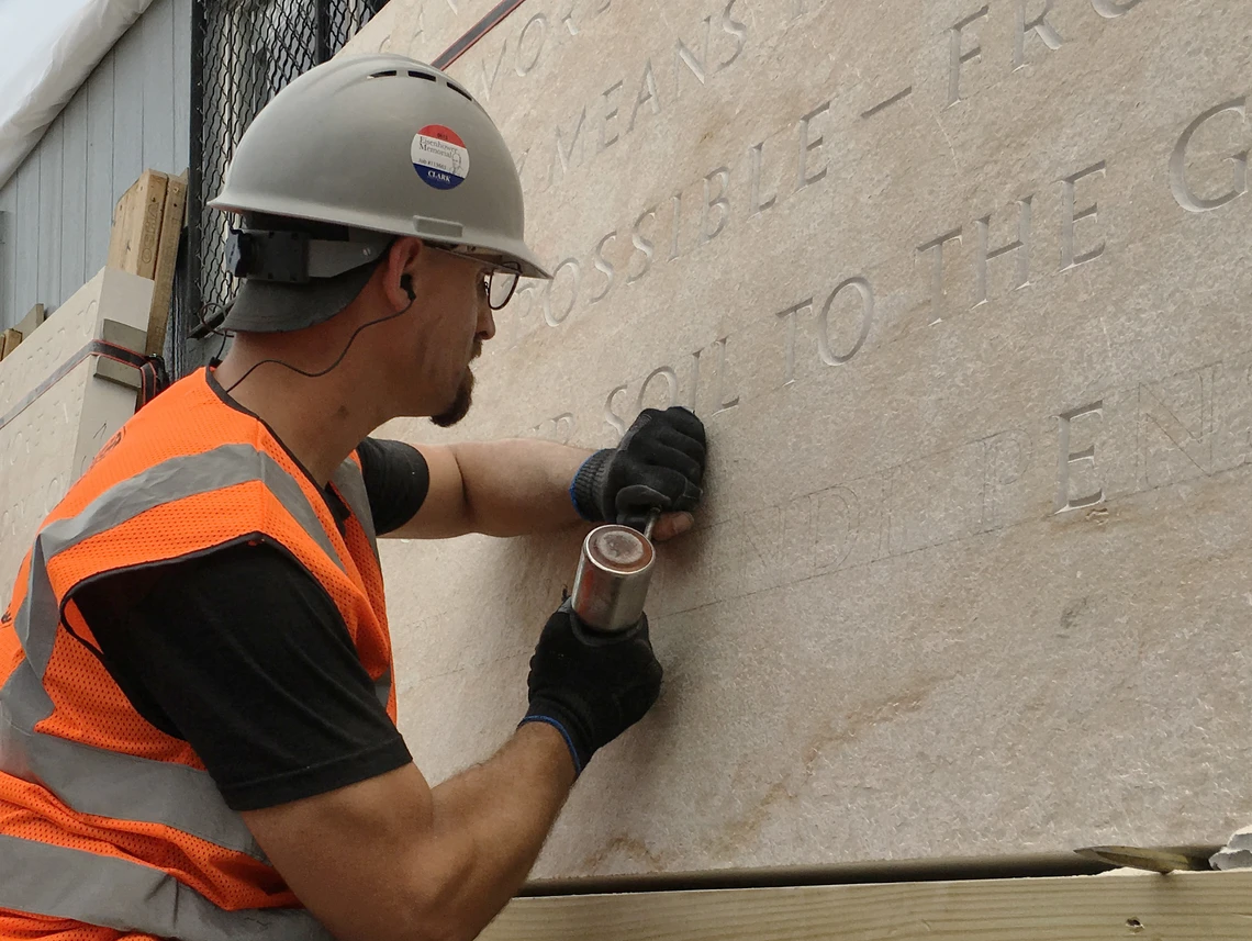 A man in a hard hat and orange safety vest works outside to carve inscriptions on a stone wall with a mallet and chisel.