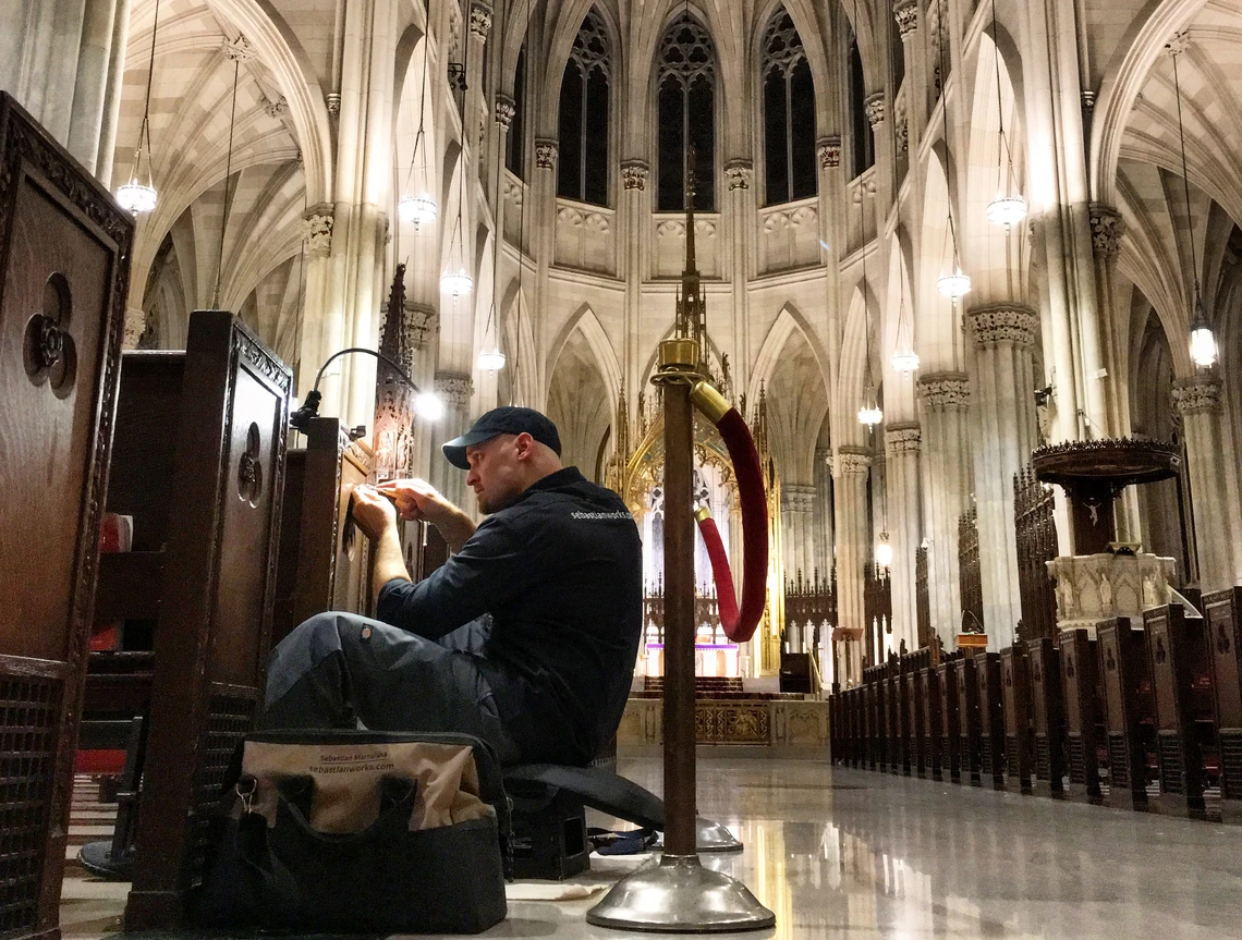 A man sitting on the floor of the center aisle in an empty church carves letters into the side of a wooden pew; a beautiful, vaulted stone ceiling is above him.