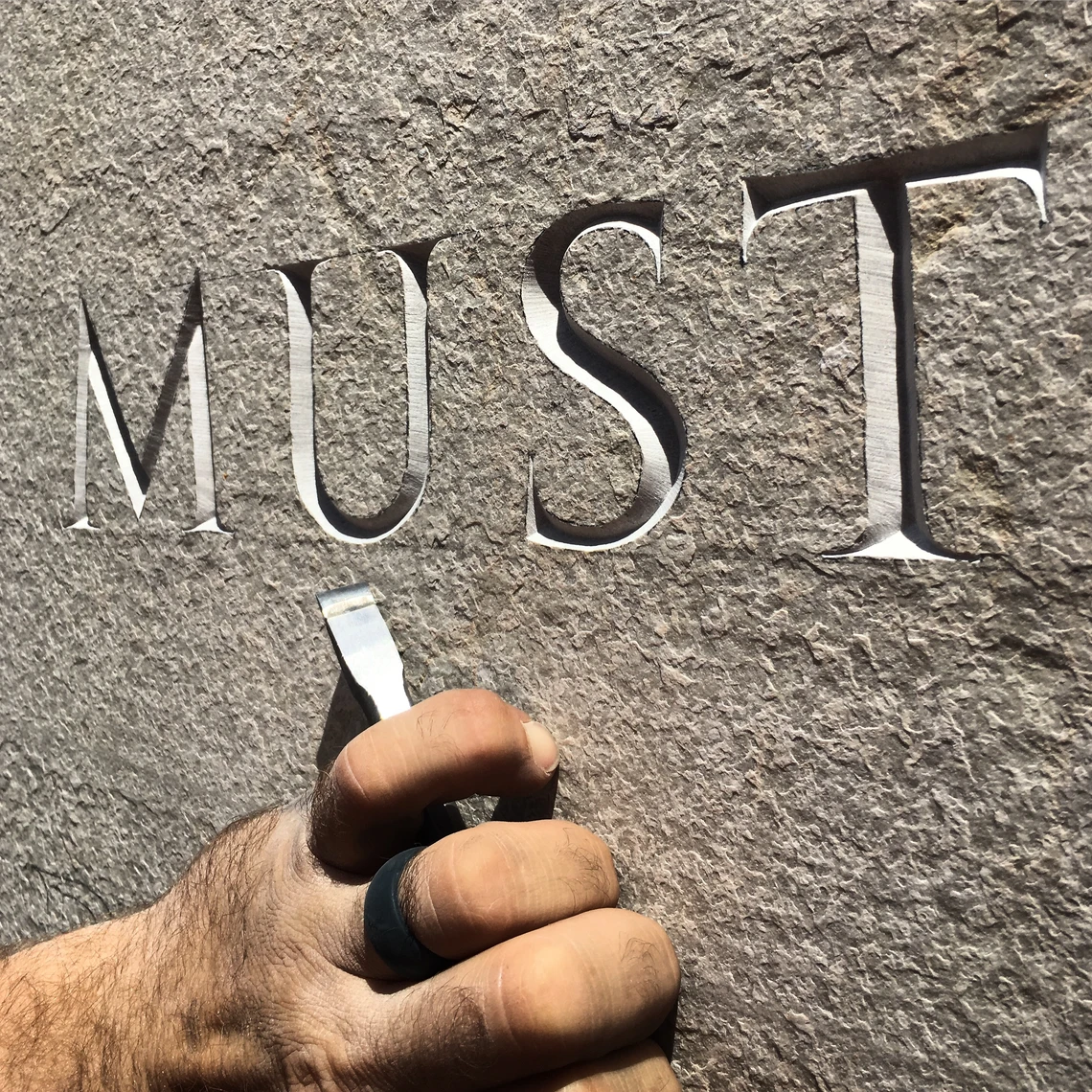 A close-up image of a man’s hand holding a chisel to carve letters in stone.