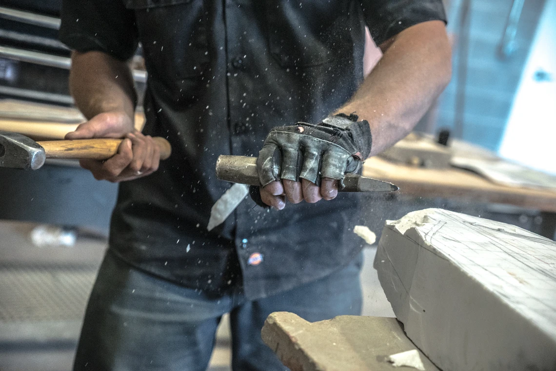 This image is a close-up of a man’s hands using a hammer to strike a thick chisel to carve a piece of marble on a workbench in a carving studio.